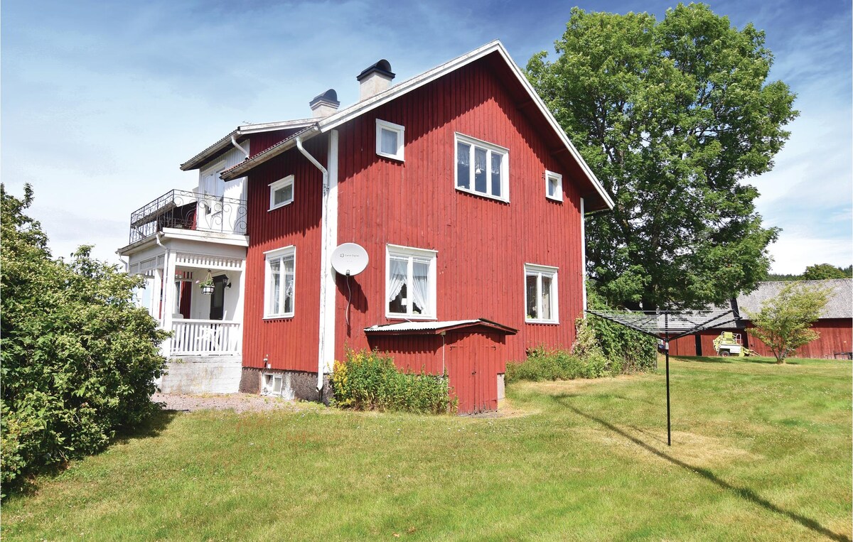 A well-maintained red wooden house is shown, featuring multiple windows and a balcony on the upper level. The surrounding yard is grassy and includes several shrubs, providing a natural setting. The image captures the essence of a charming rural home.