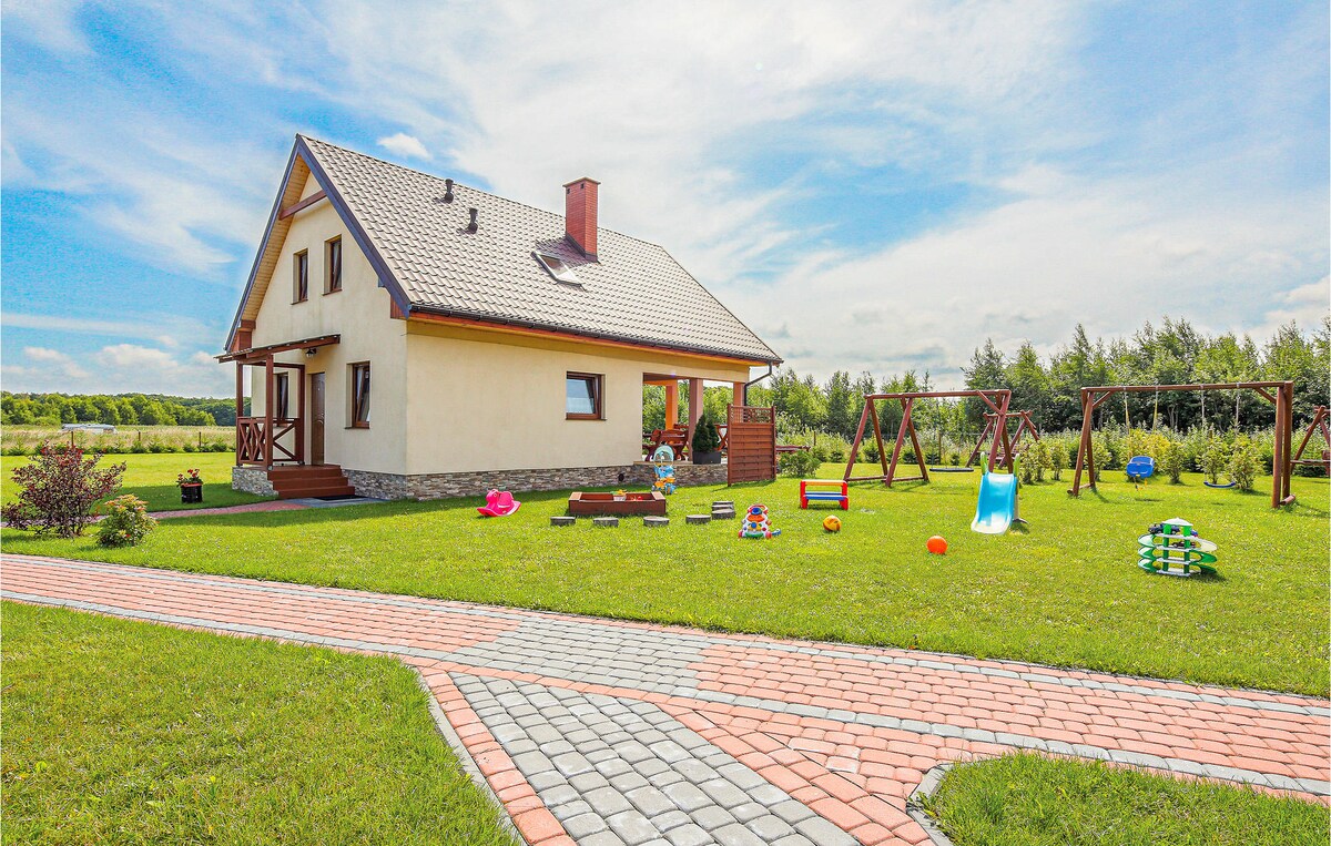 A modern cottage is set on a spacious green lawn, featuring a playground with slides and swings. A block path leads to the entrance, surrounded by trees and open fields, indicating a tranquil environment for outdoor activities.