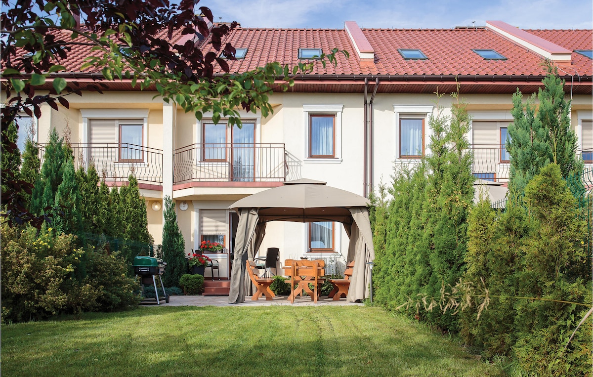 An outdoor space is featured, showcasing a gazebo with a wooden table and chairs beneath, surrounded by neatly trimmed hedges and greenery. A charcoal grill is positioned nearby, and a bright lawn provides ample space for relaxation.
