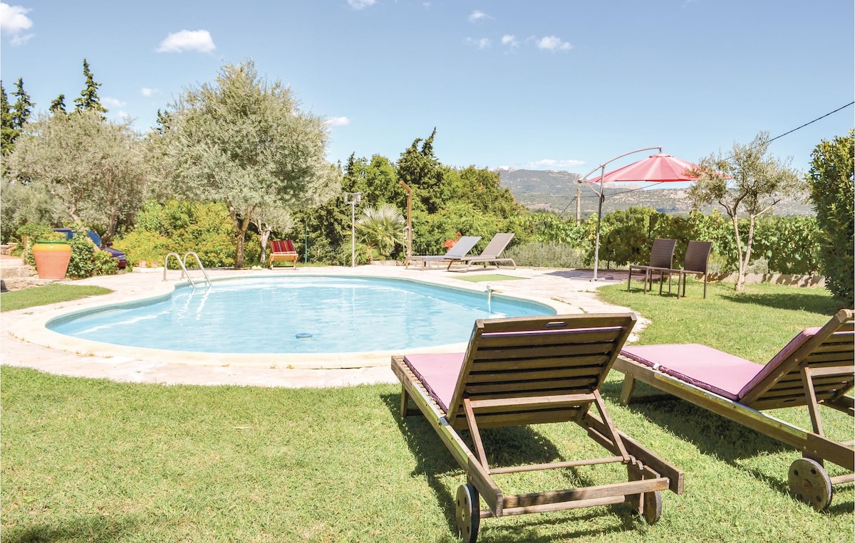 A sunlit swimming pool is surrounded by well-kept grass and trees. Two lounge chairs are positioned nearby. A red canopy provides shaded seating, while additional sun loungers are visible in the background. The landscape features lush greenery and distant hills under a clear blue sky.