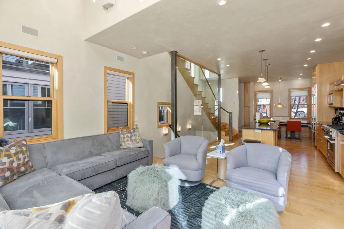 An inviting living area features a spacious gray sectional sofa paired with two light blue armchairs. Soft, round poufs add texture to the hardwood floor, while large windows provide bright light. A glass staircase leads to the upper level, with a modern kitchen visible in the background.