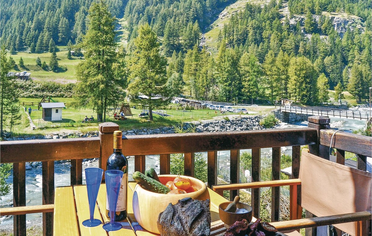 A wooden terrace is set for outdoor dining, featuring a table with a bowl of fresh vegetables, a bottle of wine, and glasses. The view includes lush green hills and a flowing river, along with a bridge connecting to the opposite bank.