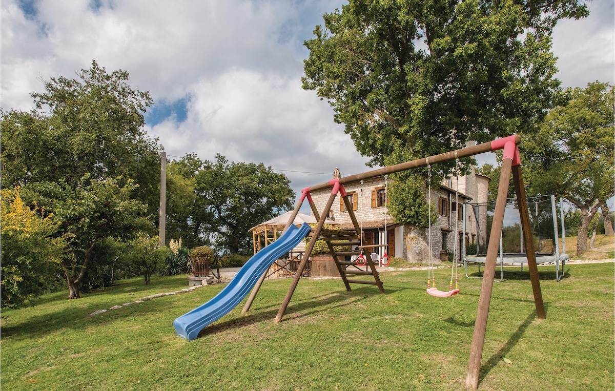 A children's play area features a blue slide and a swing set framed by green grass and trees. In the background, a rustic stone house is visible behind the play equipment, offering a welcoming and spacious environment for family activities.