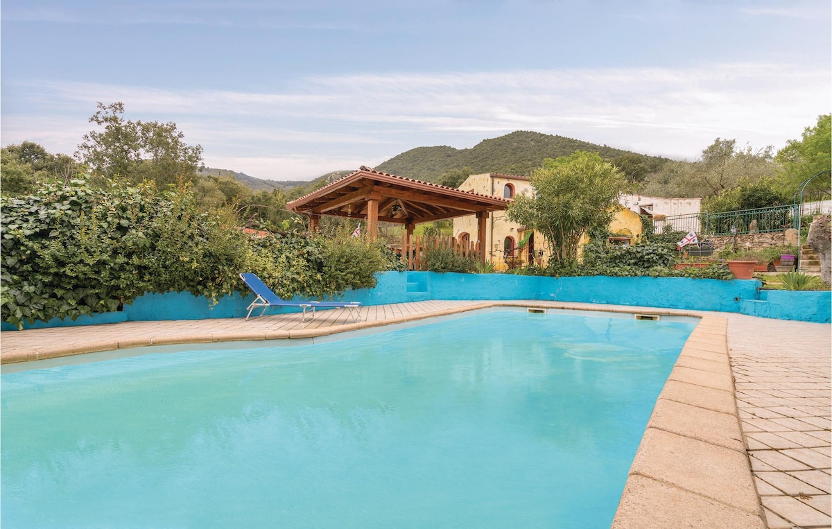 The outdoor pool is surrounded by lush greenery and a colorful, blue-tiled border. A shaded area features a wooden structure that provides shelter, and a single lounge chair sits nearby. The mountains in the background enhance the tranquil atmosphere of the space.