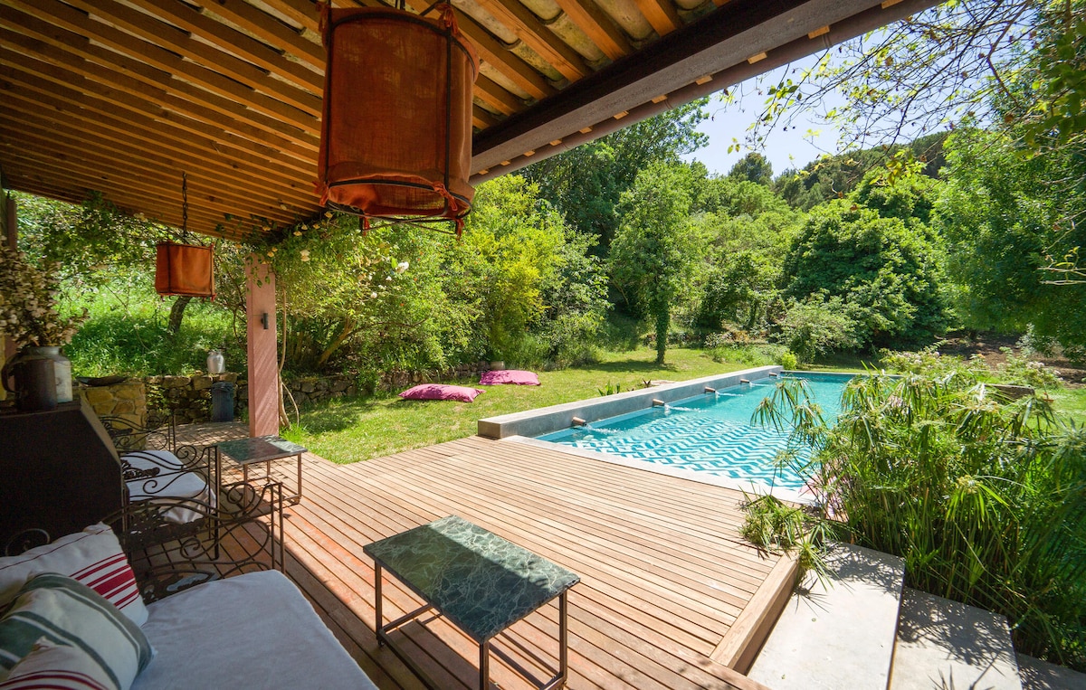 A serene outdoor area features a wooden deck surrounding a sparkling swimming pool. Sun loungers are positioned on the grass, and lush greenery borders the space, creating a tranquil retreat. Soft lighting fixtures hang from the ceiling, adding a cozy touch to the ambiance.