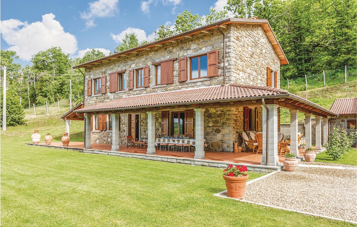 A charming Tuscan stone house is situated amidst lush greenery, featuring a spacious covered terrace supported by stone columns. Various seating areas are available on the terrace, complemented by terracotta pots with vibrant flowers. The structure's wooden shutters and tiled roof add traditional character.