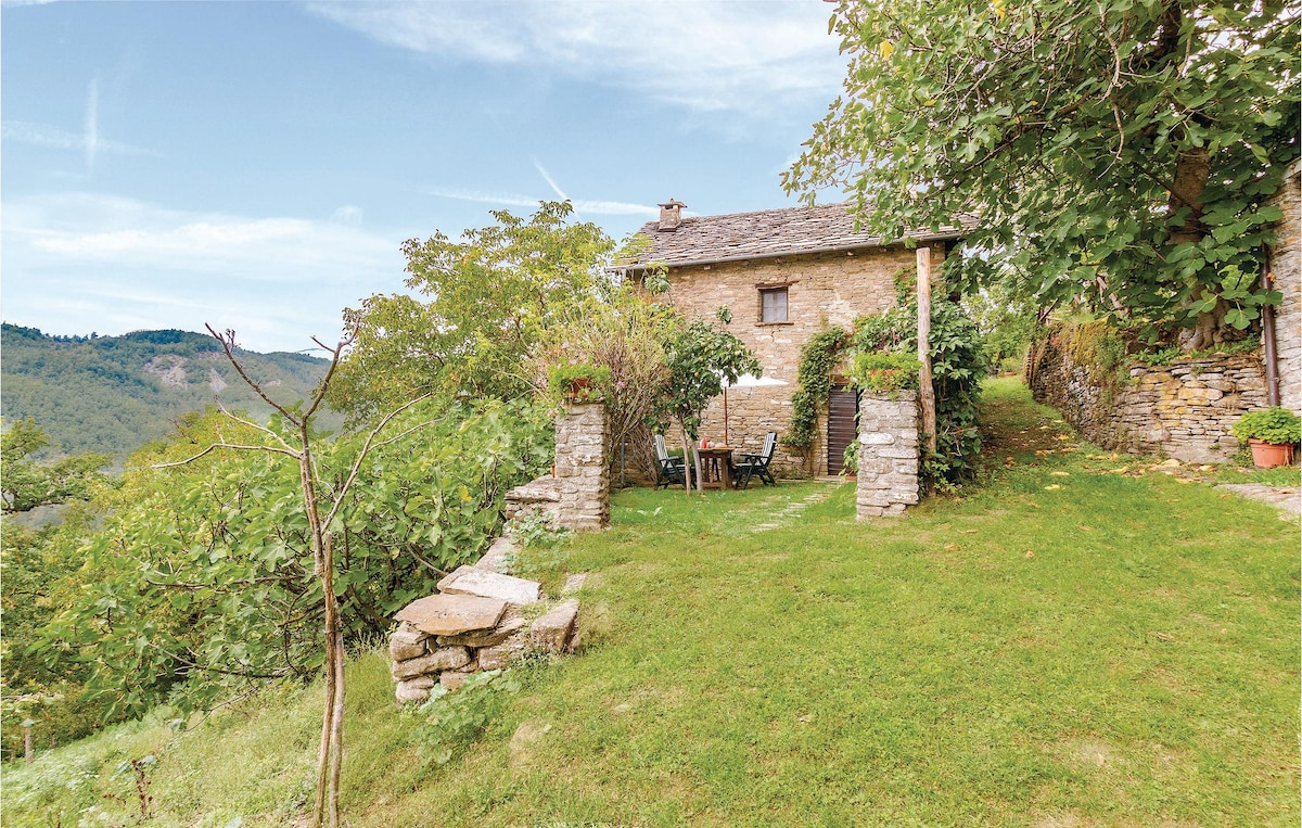 A rustic stone house sits amidst a lush, green landscape, surrounded by trees and shrubs. A small patio area with chairs is visible in front, offering a serene view of the rolling hills in the distance, complemented by blue skies.