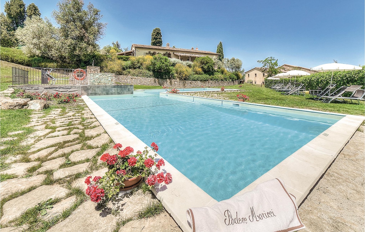 A large pool is surrounded by well-maintained stone pathways and vibrant flower beds. Sun loungers and parasols are positioned nearby, providing shaded relaxation. Lush greenery and renovated farmhouse buildings create a serene backdrop, reflecting the tranquil setting of the Tuscan countryside.