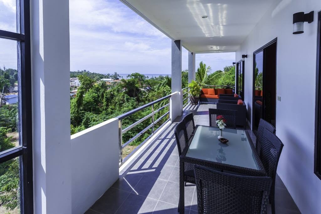 A wide terrace is seen featuring a dining table surrounded by black wicker chairs. A small floral arrangement sits on the table. An outdoor lounge area with a sofa is visible, framed by tropical greenery and views of the landscape and sea.