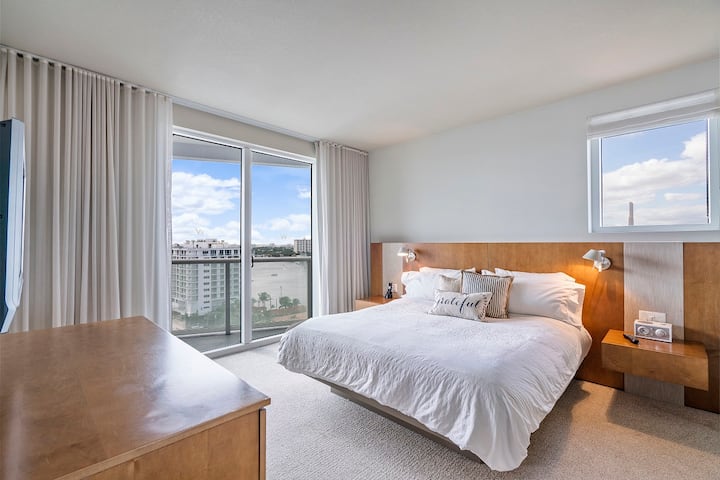 Un dormitorio moderno con una gran cama king, cabecera de madera, mesitas de noche, vestidor y TV montada en la pared. La habitación tiene una ventana grande y una ventana más pequeña, ambas con vistas a los edificios y al agua en el exterior.