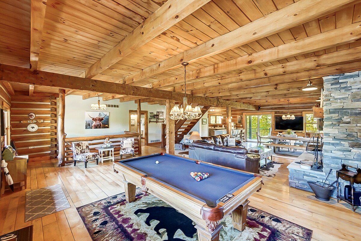 The open concept great room features rustic wooden beams and a polished wood floor, with a pool table at the center. A spacious seating area includes a dark leather sofa and two cozy chairs. Large glass doors provide access to the outdoor space, with a stone fireplace accentuating the interior.