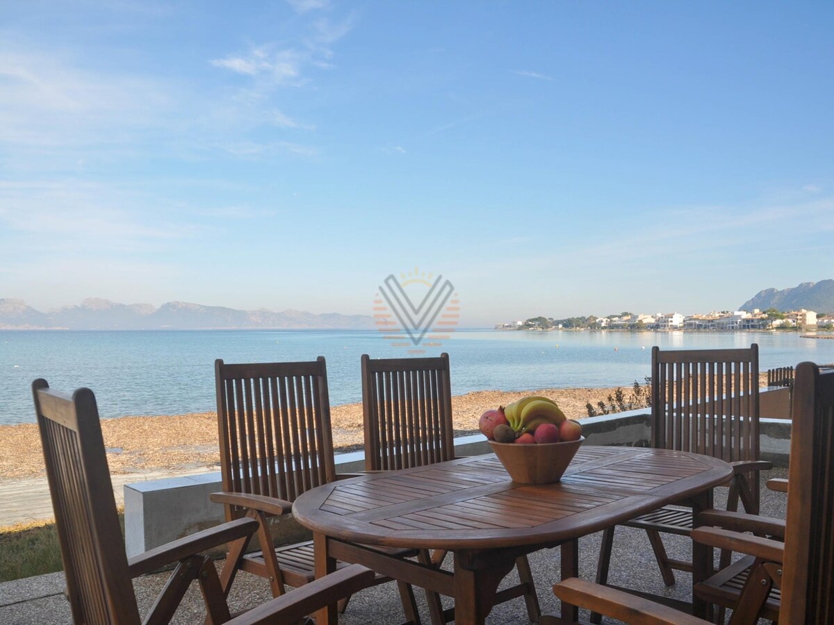 A wooden dining table with several slatted chairs is positioned on a terrace, offering views of the calm sea and distant mountains. A bowl of fresh fruit is centered on the table. The sandy beach is visible nearby, contributing to a serene coastal ambiance.