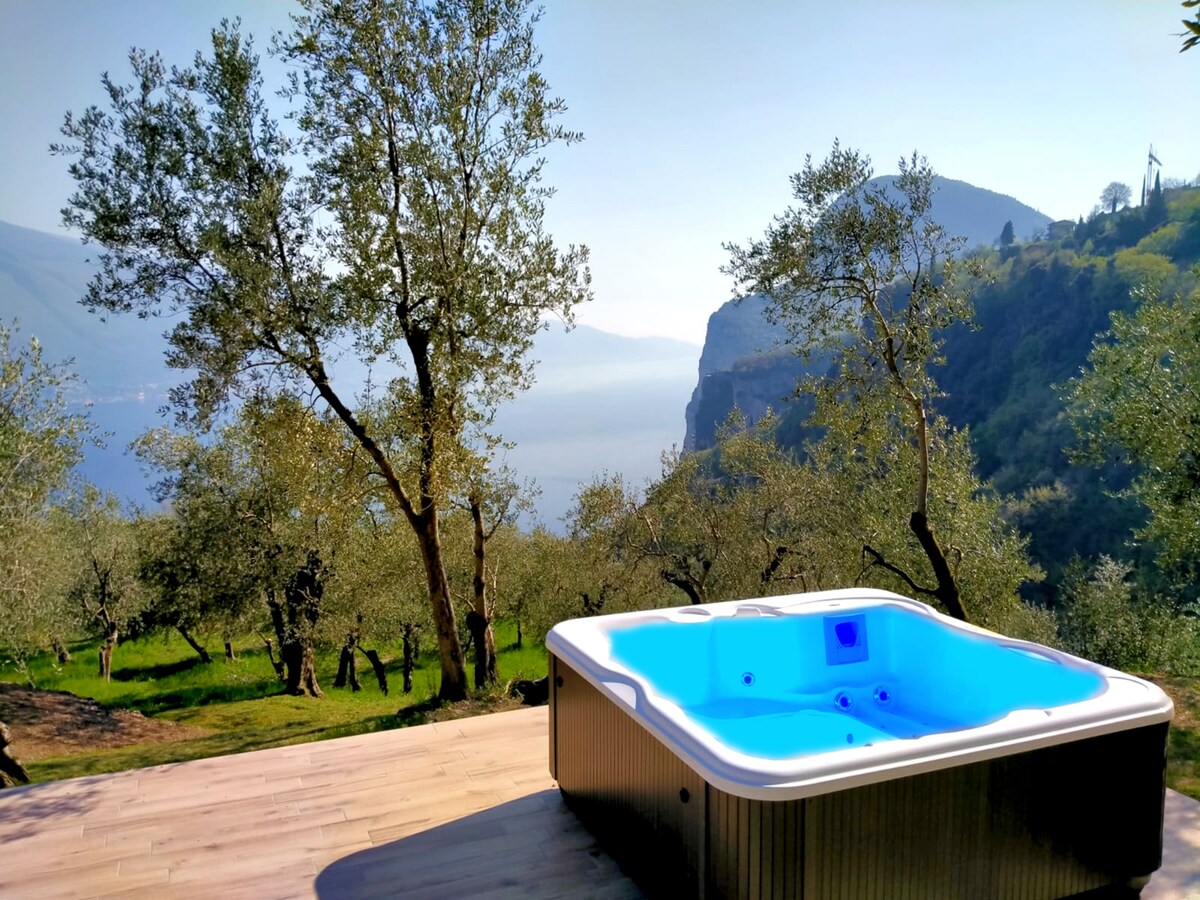 A hot tub is situated on a wooden deck, surrounded by olive trees and lush greenery. The view reveals distant mountains and a serene lake, highlighted by natural light. The calm atmosphere invites relaxation in a tranquil outdoor setting.