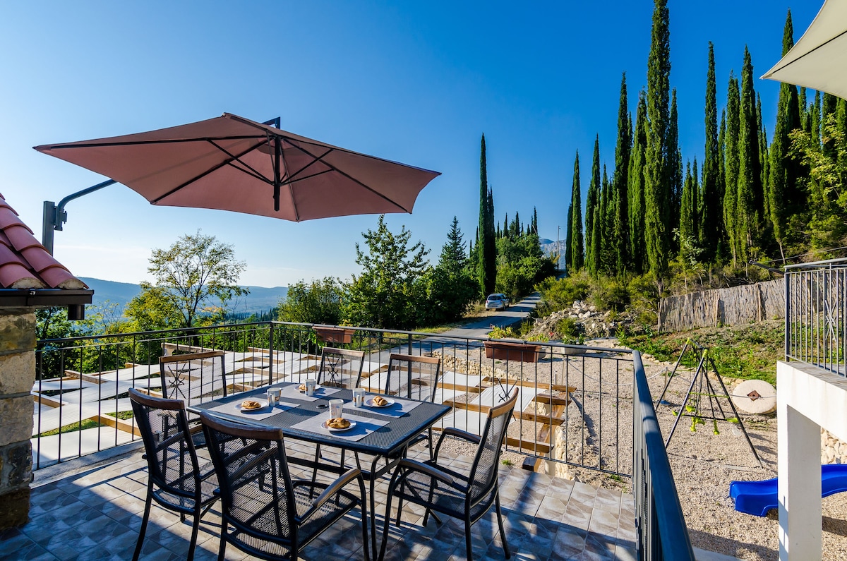 An outdoor terrace features a large dining table set for a meal, accompanied by comfortable chairs. An umbrella provides shade, while lush cypress trees and greenery surround the area, enhancing the serene atmosphere. A children's playground is visible in the background, contributing to the family-friendly environment.
