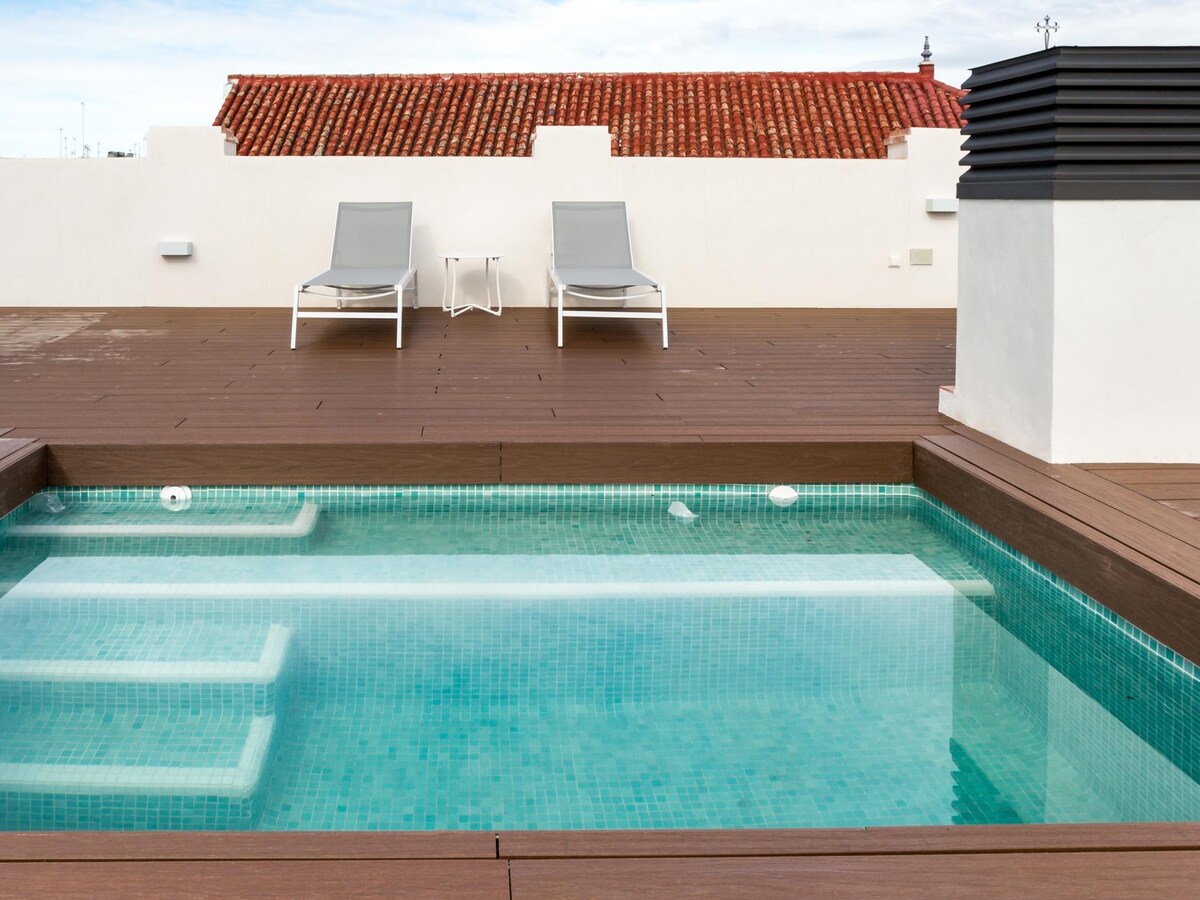 A small shared pool is situated on a wooden deck, surrounded by neutral-toned lounge chairs. The area offers a relaxed atmosphere with a clear view of the sky above, creating a serene space for guests to enjoy.