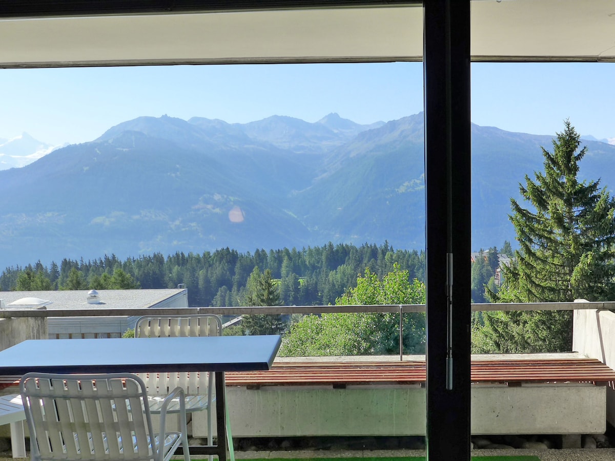 A spacious balcony offers a clear view of the surrounding mountains and lush countryside. Outdoor furniture, including a table and benches, provides opportunities for relaxation while enjoying the natural scenery. The glass doors allow natural light to enter the living space.