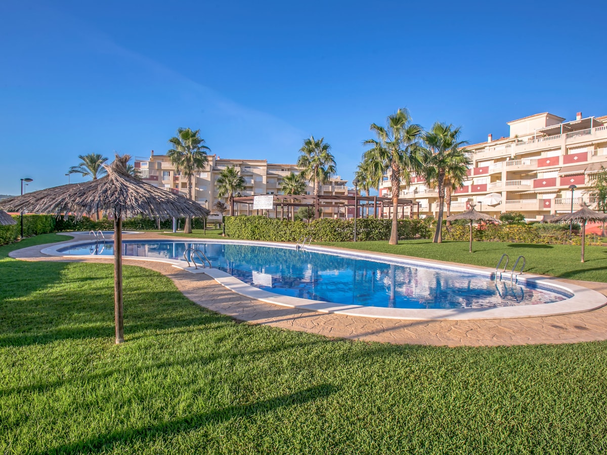 A kidney-shaped swimming pool is surrounded by well-maintained grass and palm trees, providing a serene outdoor setting. The pool area includes sun loungers and a thatched-roof shade structure, enhancing the space for relaxation and enjoyment.