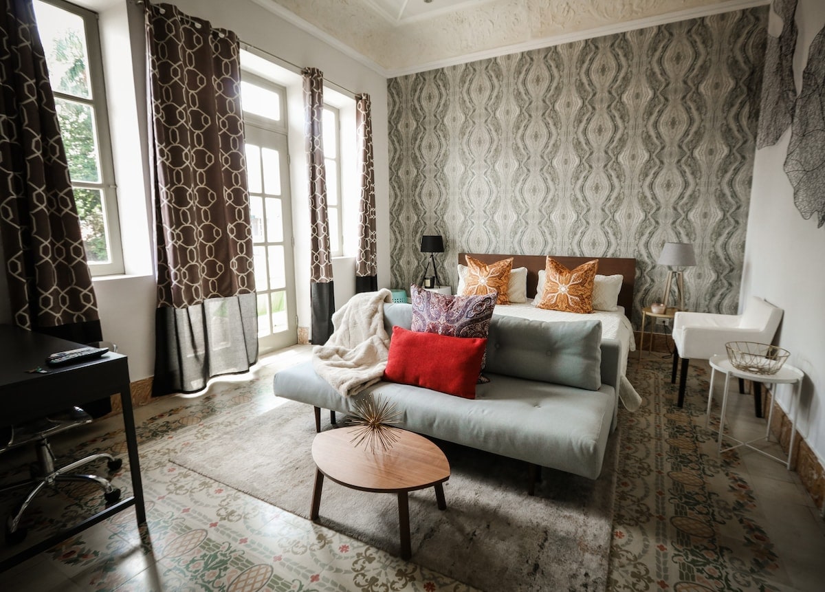 A stylish room features a spacious sleeping area with a comfortable bed dressed in tasteful bedding. A cozy seating area is highlighted by colorful throw pillows and a coffee table. Natural light enters through large doors, framed by elegant curtains, enhancing the inviting decor.