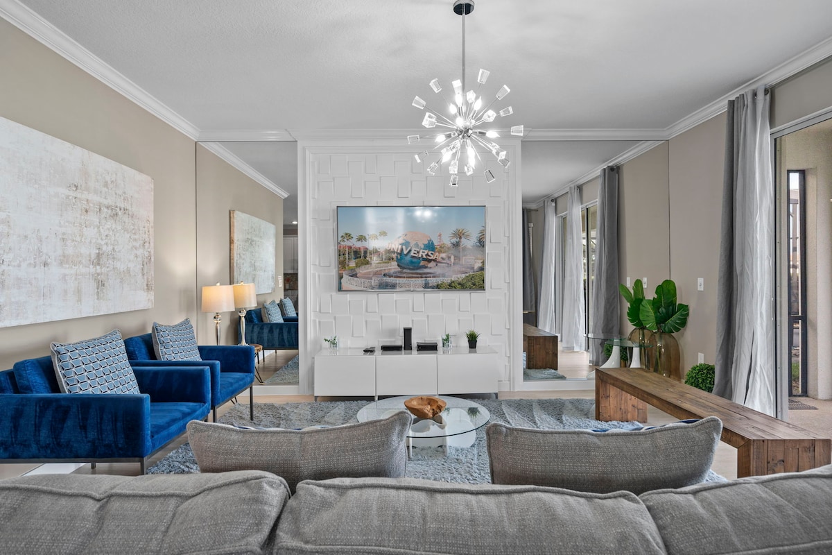 A spacious living room features a comfortable grey couch facing a modern entertainment unit with a large screen. Two blue chairs are positioned nearby, complementing the light-colored walls. Natural light filters through sheer curtains, adding warmth to the space.