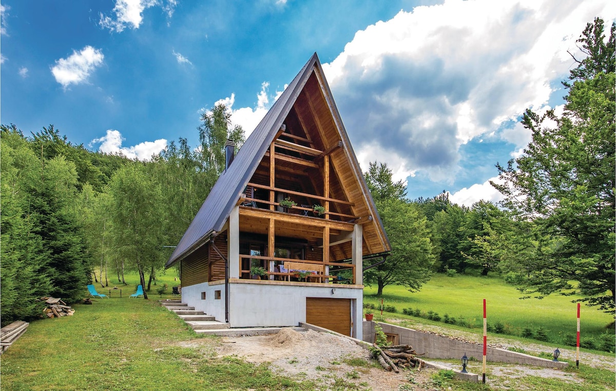 The cottage features a distinctive a-frame structure with a sloped roof, surrounded by lush greenery. Outdoor seating areas and wooden accents are visible on the deck. A gravel path leads to the entrance, set against a backdrop of rolling hills and a bright blue sky.