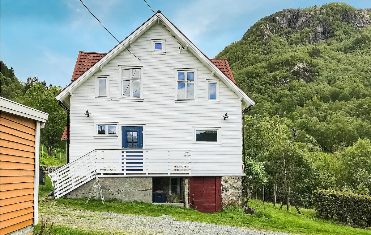 The exterior of a charming two-story cottage is shown, featuring white wooden siding and red roof tiles. A spacious deck leads to the entrance, while surrounding greenery and rolling hills create a natural backdrop. Windows allow ample light to enter the home.