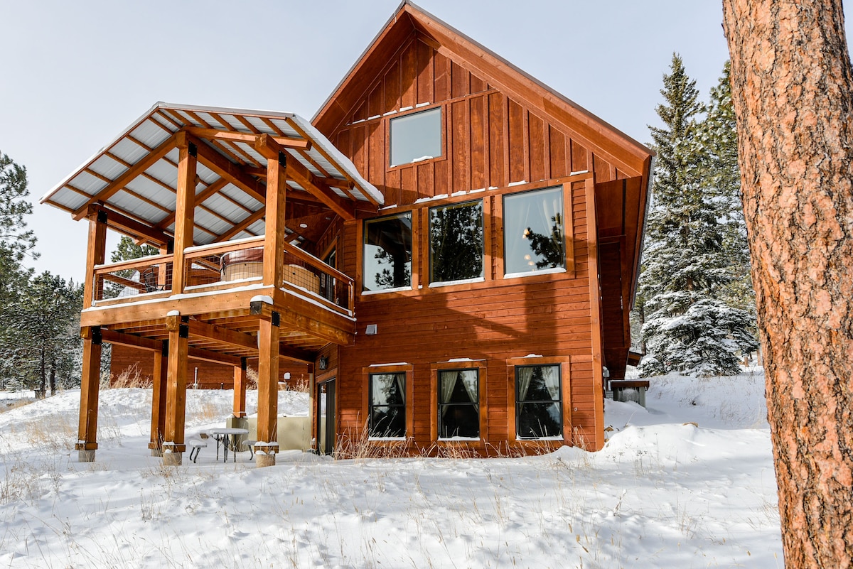 The exterior of the modern cabin showcases wooden construction with large windows, allowing natural light to fill the interior. A covered deck extends from the upper level, supported by log columns. Surrounding snowy terrain and evergreen trees add to the serene setting.