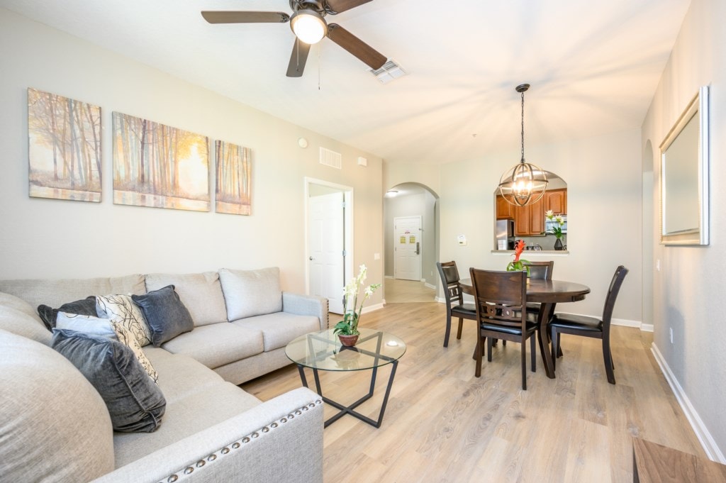 A comfortable living area features a spacious gray sectional sofa with decorative pillows, positioned beside a glass coffee table. A round dining table with dark wooden chairs is set nearby. The walls are adorned with a serene nature-themed artwork, enhancing the inviting ambiance.