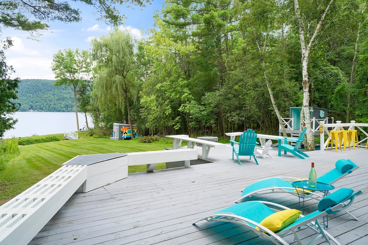 A spacious deck is adorned with colorful lounge chairs and tables, offering a seating area overlooking the lush greenery and tranquil waters. The well-maintained lawn extends towards the lake, with trees providing shade and a serene backdrop for outdoor relaxation.