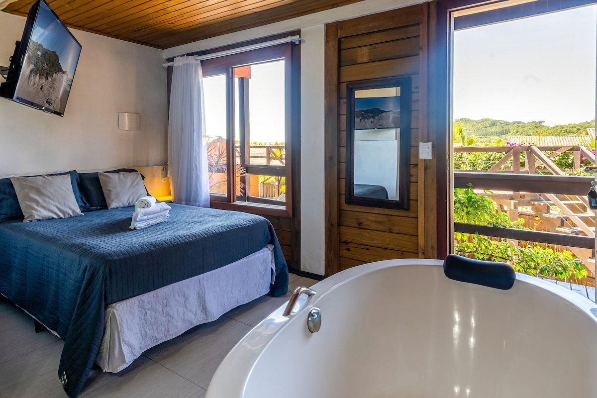 A spacious suite features a large bed with crisp linens, accompanied by a comfortable armchair. Large windows provide natural light and views of the lush surroundings. A modern bathtub is positioned nearby, enhancing the room's functionality and relaxation options.