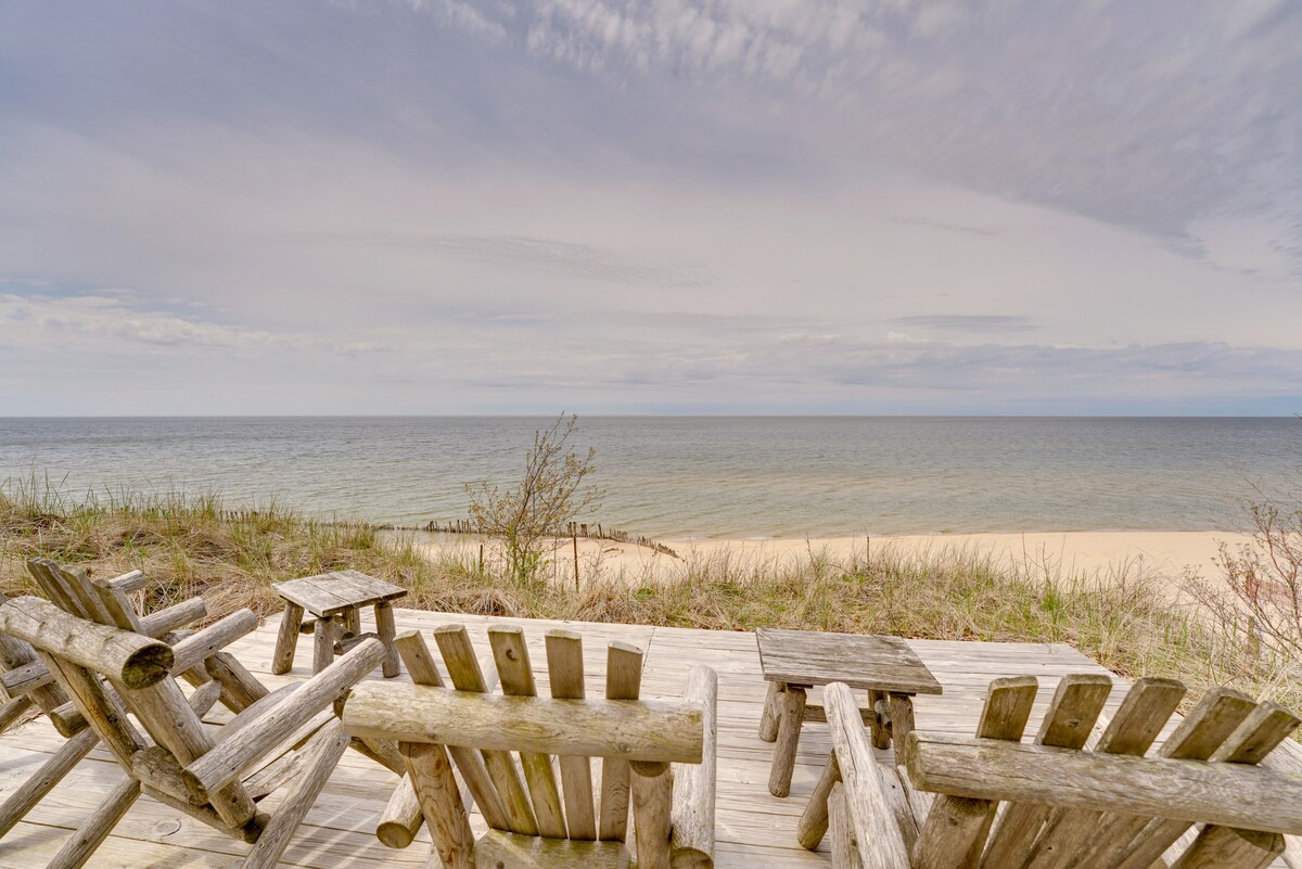 A wooden deck features several rustic chairs arranged in a circle, offering views of the tranquil lake and sandy beach. The sky displays soft clouds, while grass and shrubbery adorn the edges of the deck, blending with the natural beach landscape.