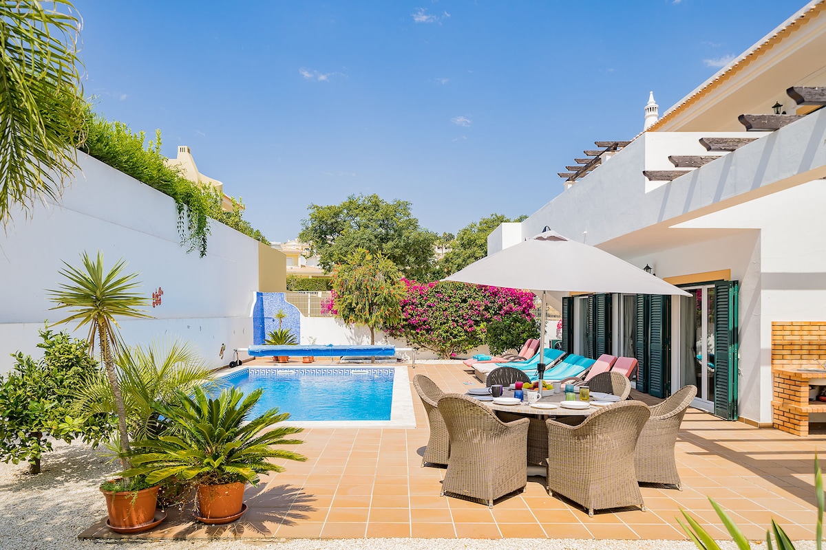 The outdoor area features a private swimming pool surrounded by a tiled patio. Comfortable seating is arranged around a dining table, with potted plants adding greenery. A large umbrella provides shade. Vibrant flowering bushes enhance the tranquil space, inviting relaxation.