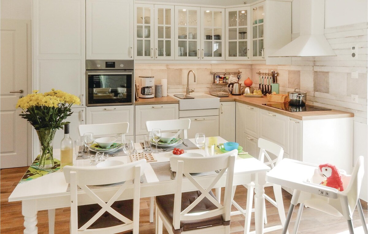 A well-equipped kitchen features white cabinetry with glass doors, showcasing kitchenware. A dining table is set for six, complemented by white chairs. Fresh flowers in a vase add a touch of color to the space, illuminated by natural light from a window.