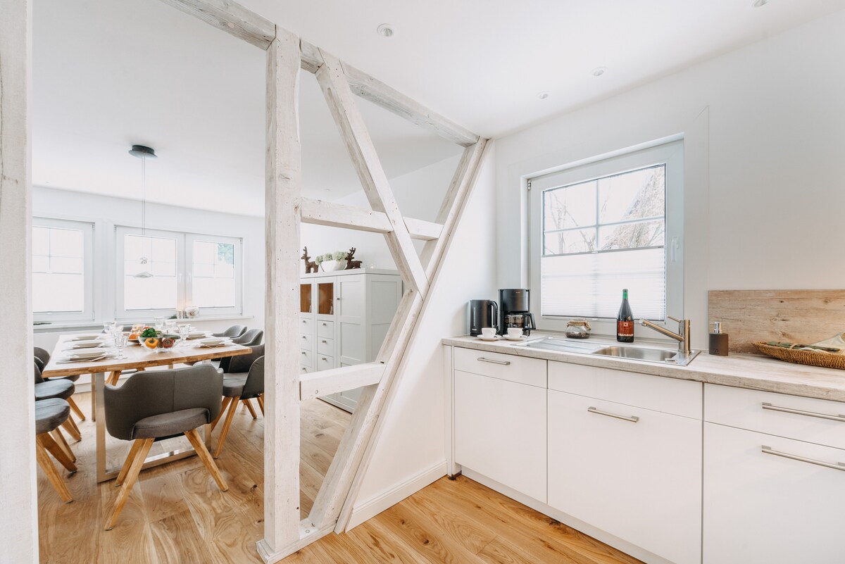A modern kitchen is depicted, featuring sleek white cabinets, a countertop with a sink, and appliances including a coffee maker. A large dining area with a wooden table and ergonomic chairs is visible through an adjacent doorway, illuminated by natural light from the window.