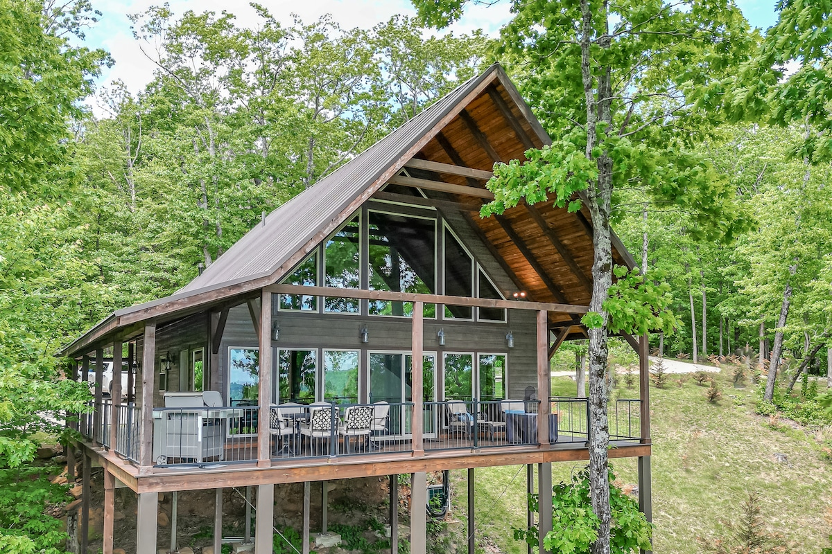 A treetop cabin is set among lush greenery, featuring a striking A-frame design. Large windows provide ample natural light and showcase expansive views. The covered deck is furnished with a dining table and seating, creating an inviting outdoor space.