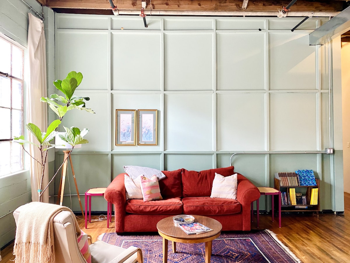A bright living area showcases a comfortable red couch placed against a pale green panel wall. Two framed artworks adorn the wall above a small round table, while natural light filters through the windows. A fiddle leaf fig plant adds a touch of greenery to the space.