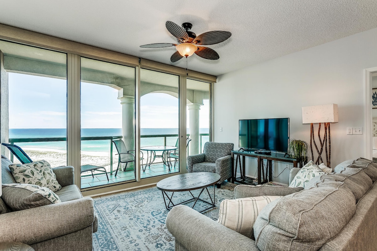 A spacious living area is depicted with large glass doors allowing natural light and views of the Gulf of Mexico. Comfortable seating is arranged around a round coffee table. A ceiling fan enhances airflow, while a flat-screen television is positioned for easy viewing.