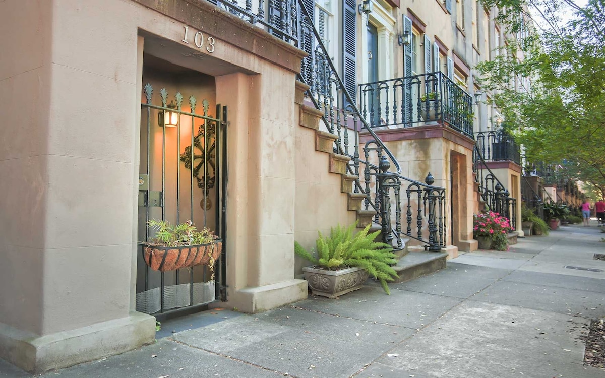 The entrance to the garden apartment is framed by a decorative wrought iron gate. Lush greenery and colorful flowers enhance the inviting exterior. A set of stairs leads to the upper levels of the building, with a charming facade characterized by classic architectural details.