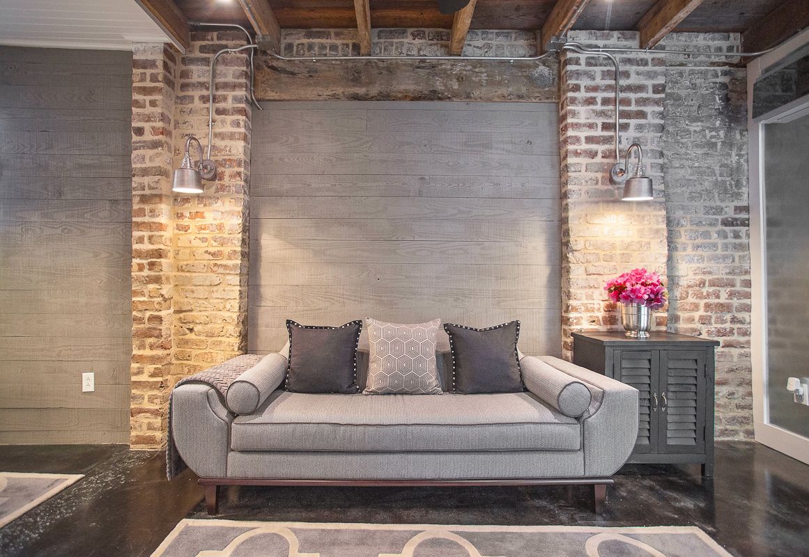 A stylish living area features a grey couch, adorned with decorative pillows, against a backdrop of alternating exposed brick and grey wood panel walls. Subtle lighting is provided by wall-mounted sconces, complementing the warm tones of the space.