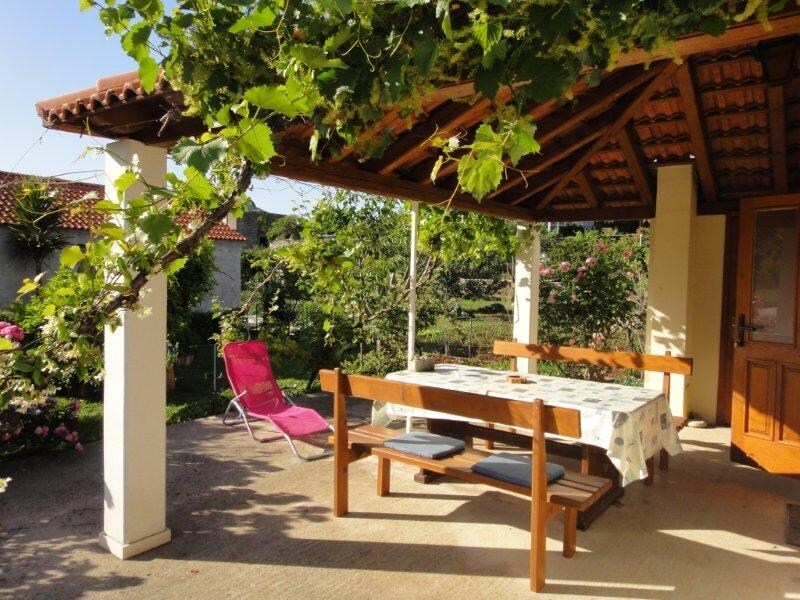 A shaded terrace features a wooden dining table with benches, surrounded by vibrant greenery and blooming flowers. A reclining lounge chair is positioned nearby, inviting relaxation in a tranquil outdoor setting. The structure's roof is supported by sturdy columns, enhancing the open atmosphere.