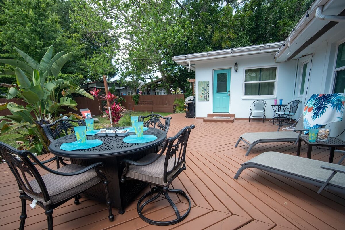 A spacious wooden deck is furnished with a round dining table surrounded by chairs and lounge chairs. Lush plants and vibrant flowers enhance the outdoor area, providing a relaxing setting. The white cottage exterior is visible, with a welcoming entrance nearby.