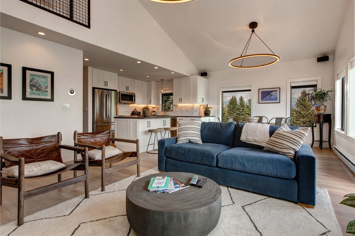 An open living area showcases a blue sectional sofa adorned with striped pillows, paired with two wooden chairs. A circular coffee table sits in front, and natural light fills the space through large windows, highlighting the contemporary kitchen in the background.