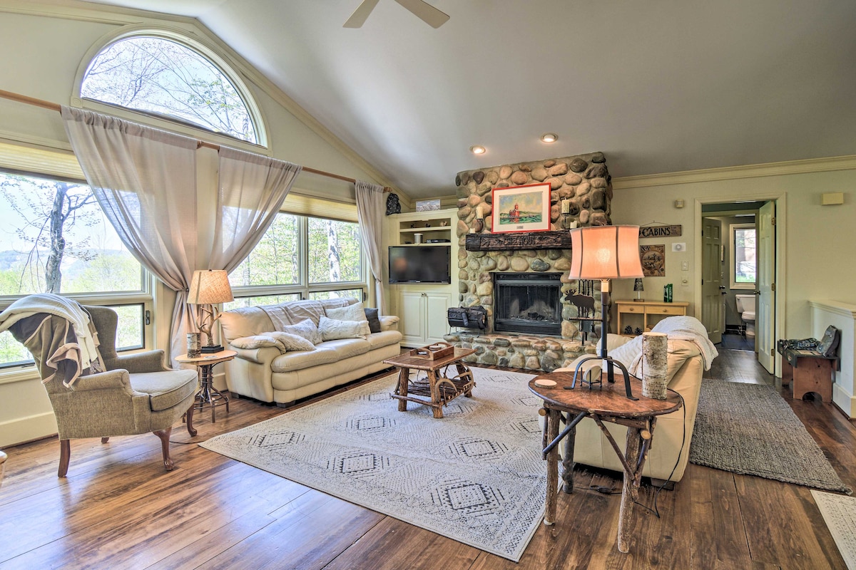 A spacious living area features a stone fireplace as a focal point, surrounded by large windows offering views of the outdoors. Comfortable seating is arranged around a central area rug, and natural light streams in through the high, arched window.