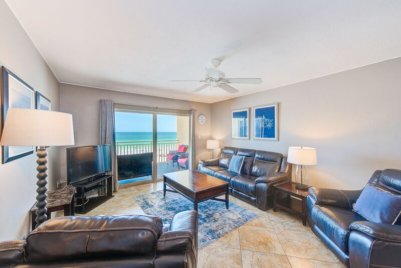 A living area is presented with leather seating arrangements and a wooden coffee table. A television is positioned against the wall, and large sliding glass doors open to a private balcony, offering views of the beachfront. Natural light fills the room through the doors.
