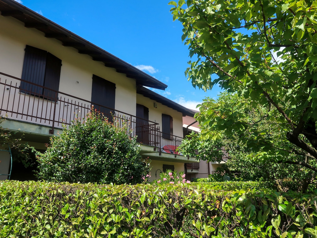The exterior of the terraced house is shown, surrounded by lush greenery and a trimmed hedge. Two balconies are visible, adorned with plants. The building features muted tones with wooden elements, complementing the clear blue sky above.