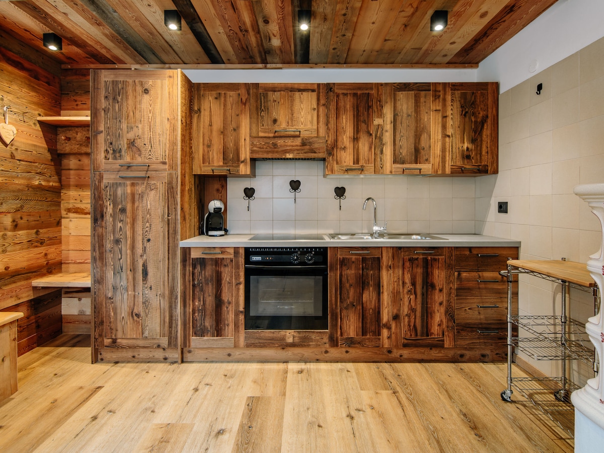 The kitchenette features wooden cabinetry and a light countertop, equipped with an oven, induction hot plates, and a coffee machine. A sleek metal cart sits beside the countertop, while warm wooden flooring enhances the space's inviting ambience.