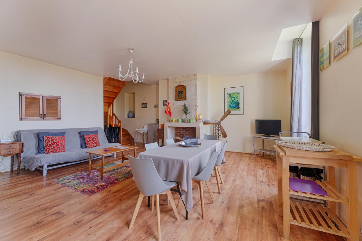 An open living space features a dining table set for six and a cozy sitting area with a grey sofa. A wooden staircase leads to an upper level, while natural light enters through a window and a doorway. A television is visible in the corner.