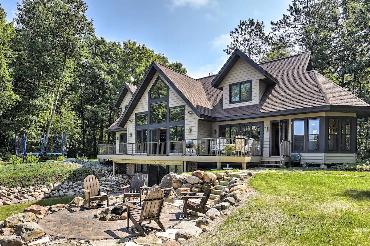 The exterior of a spacious home is depicted, featuring large floor-to-ceiling windows that allow natural light to brighten the interior. A fire pit surrounded by seating is located in the foreground, with a well-maintained lawn and a trampoline visible in the background.