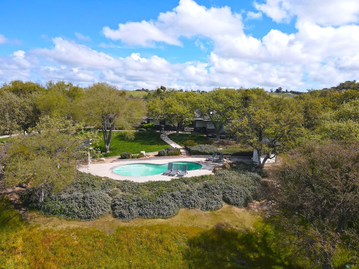 Aerial view of a serene outdoor space featuring a circular swimming pool surrounded by lush greenery. The landscaped area includes walking paths and scattered trees under a blue sky with clouds, providing a tranquil setting.