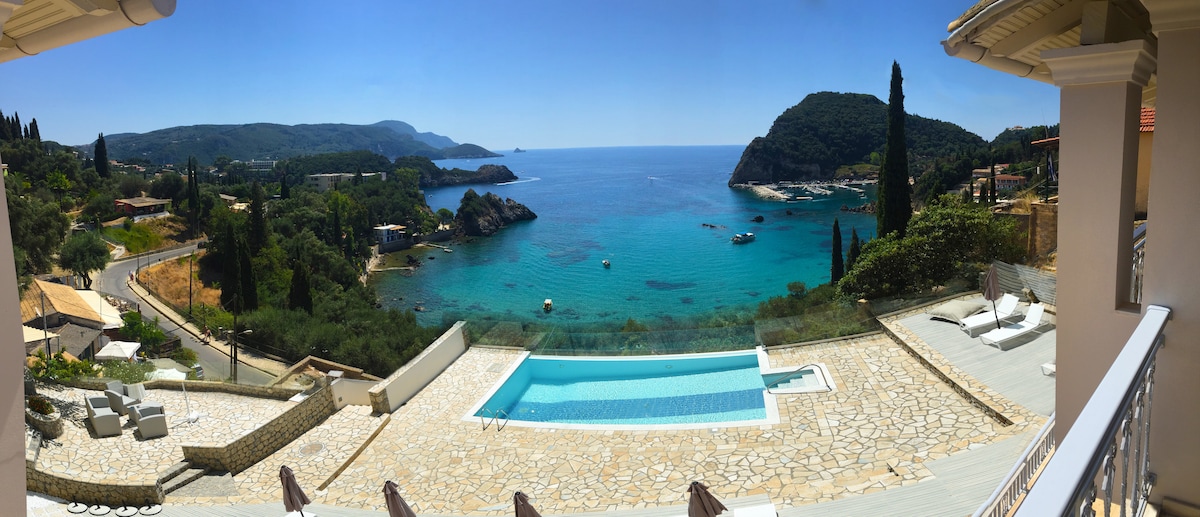 A panoramic view reveals a sparkling blue sea surrounded by lush green hills. A clear swimming pool is positioned in the foreground, while sun loungers are placed beside it. The coastline features a blend of rocky and sandy areas, creating a serene coastal scene.