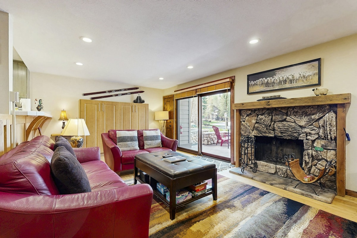 A welcoming living area features comfortable leather seating arranged around a central coffee table. A rustic stone fireplace adds warmth to the space. Large sliding glass doors open to a view of the outdoor area, allowing natural light to brighten the room.